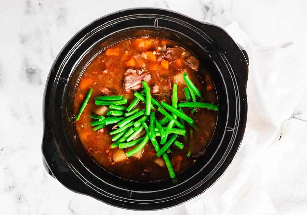 Fresh green beans added to the slow cooker with simmering beef stew, ready to be cooked.