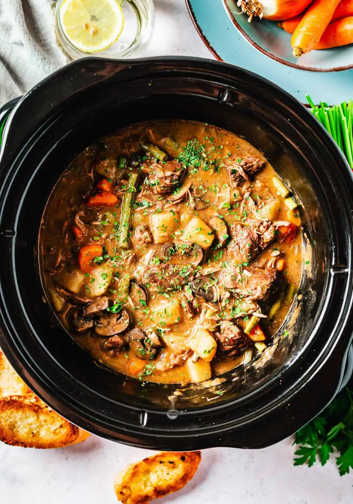 Top-down view of a slow-cooked beef stew in a crock pot, featuring mushrooms, carrots, green beans, and potatoes, garnished with chopped parsley.