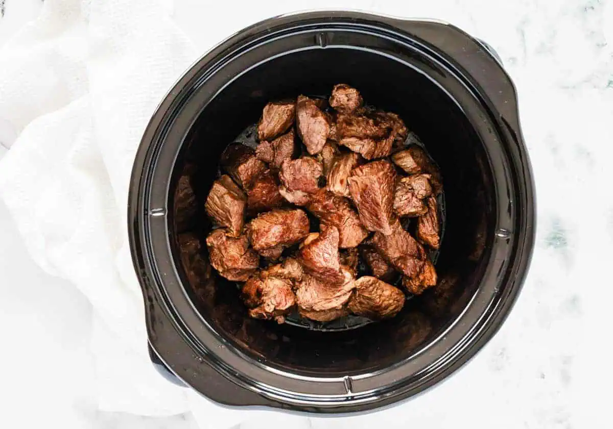 Seared beef chunks placed in the bottom of a black slow cooker.