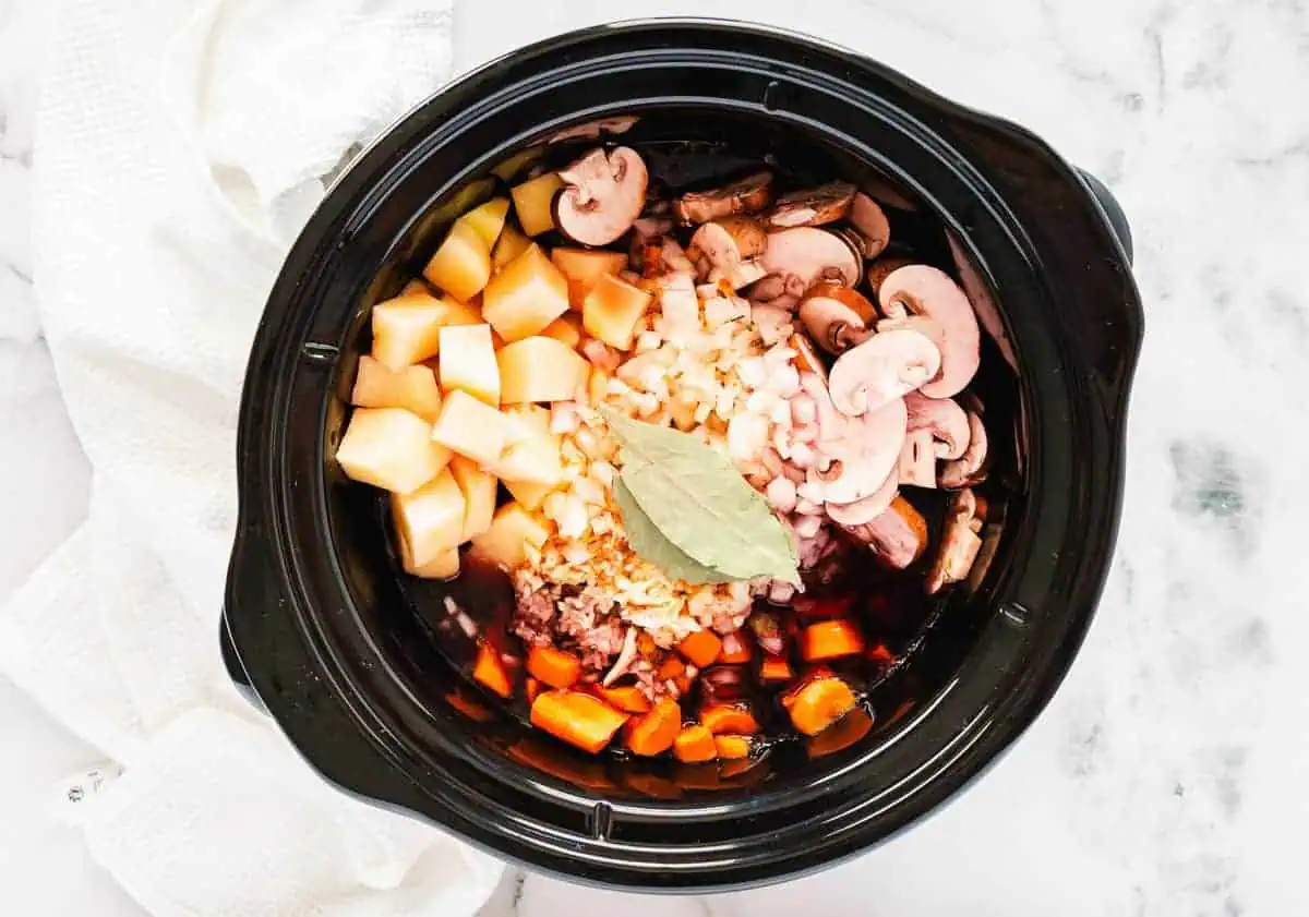 A slow cooker filled with vegetables, beef, and bay leaves, ready to start cooking.