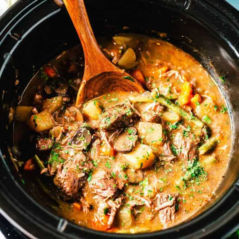 A close-up of a hearty beef stew with potatoes, carrots, mushrooms, and green beans in a slow cooker, being stirred with a wooden spoon.