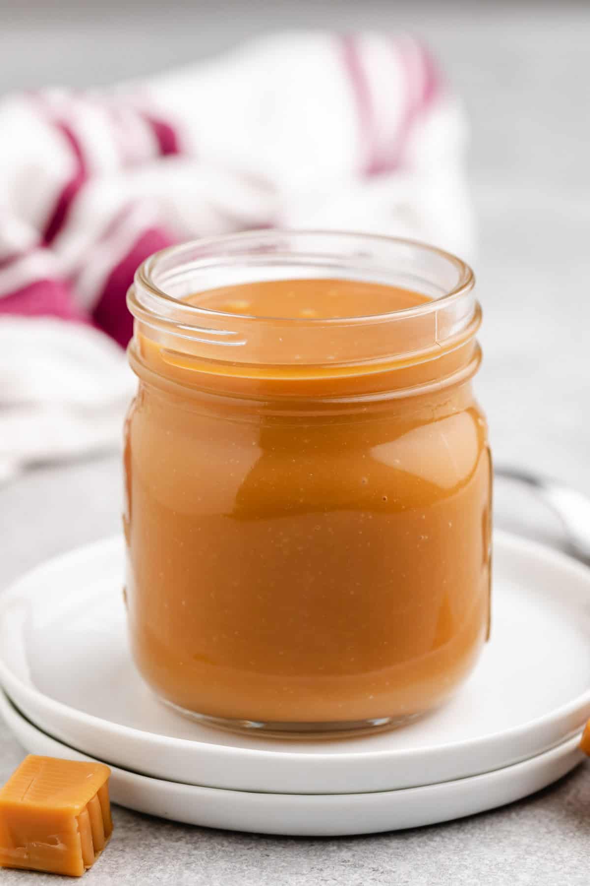 A jar of thick, creamy caramel placed on a white plate with caramel pieces beside it.