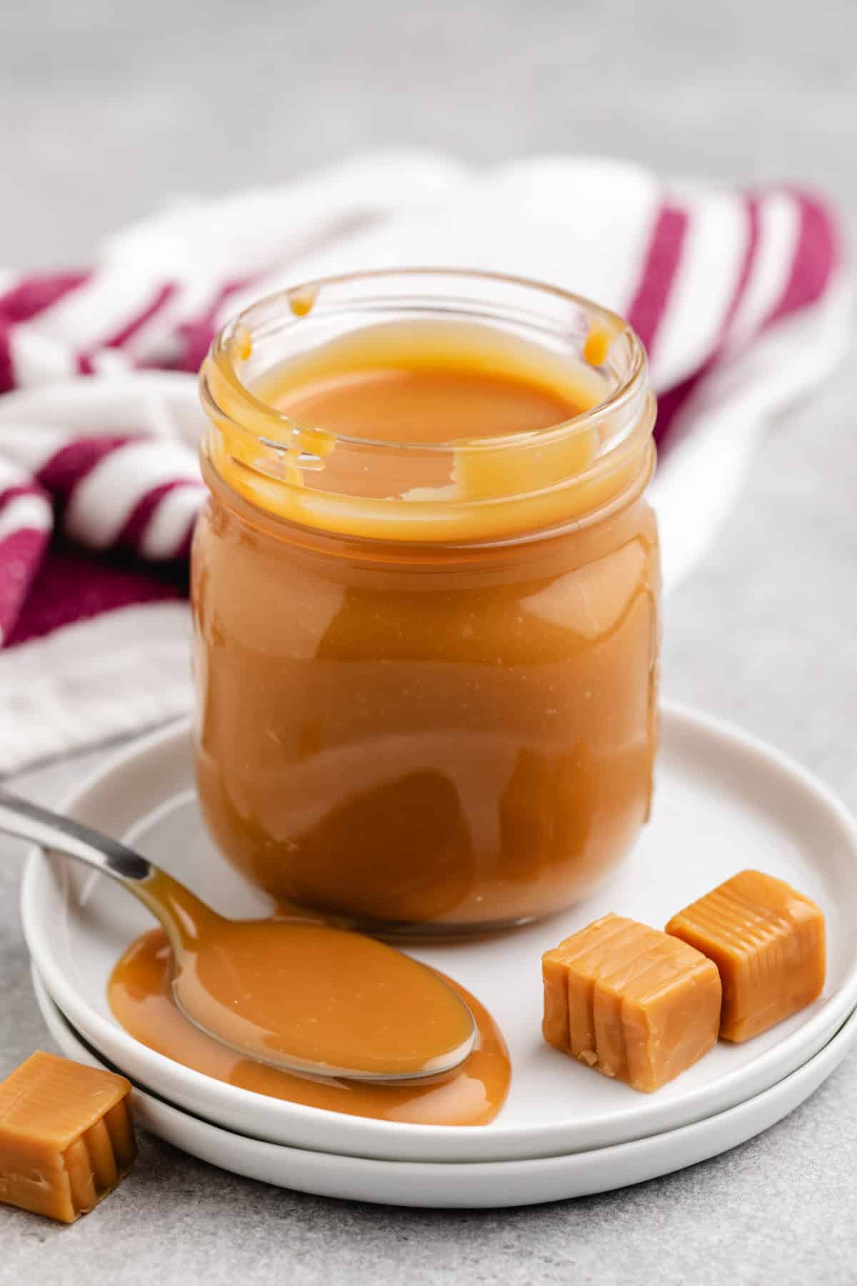 A jar of creamy caramel on a white plate with caramel squares nearby.