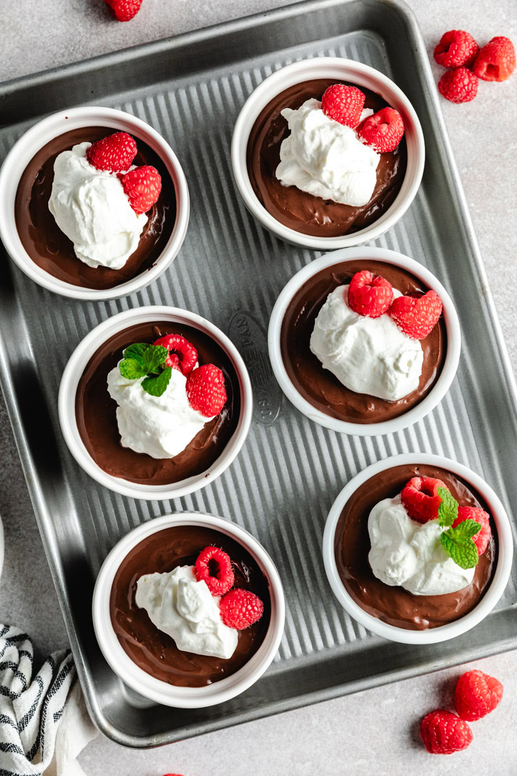 Six ramekins filled with chocolate pudding, topped with whipped cream and fresh raspberries.