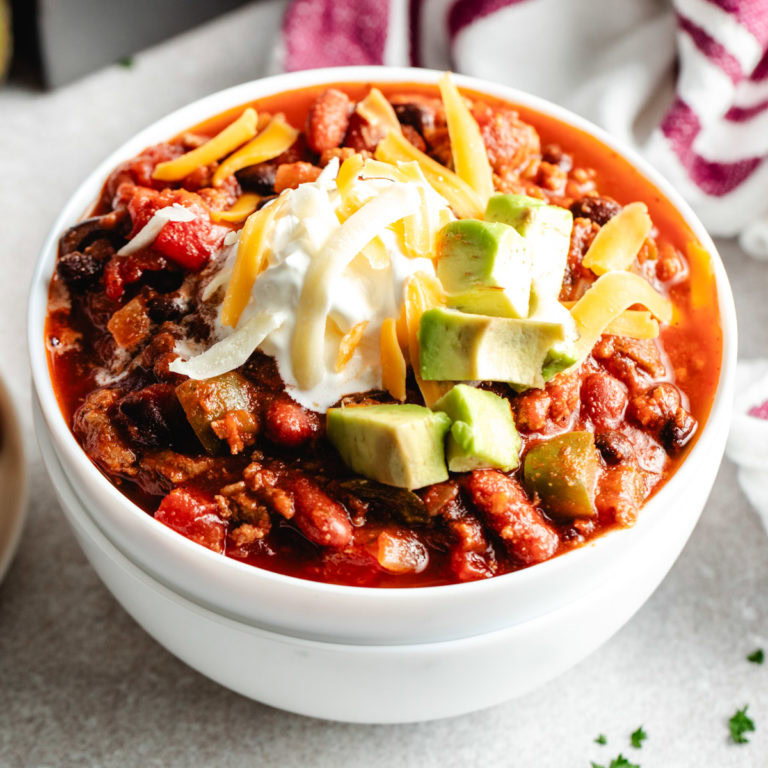 A bowl of turkey chili with a spoon, surrounded by cornbread and fresh avocado.