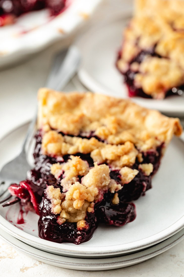 A slice of cherry pie with a golden brown topping on a stack of white plates.