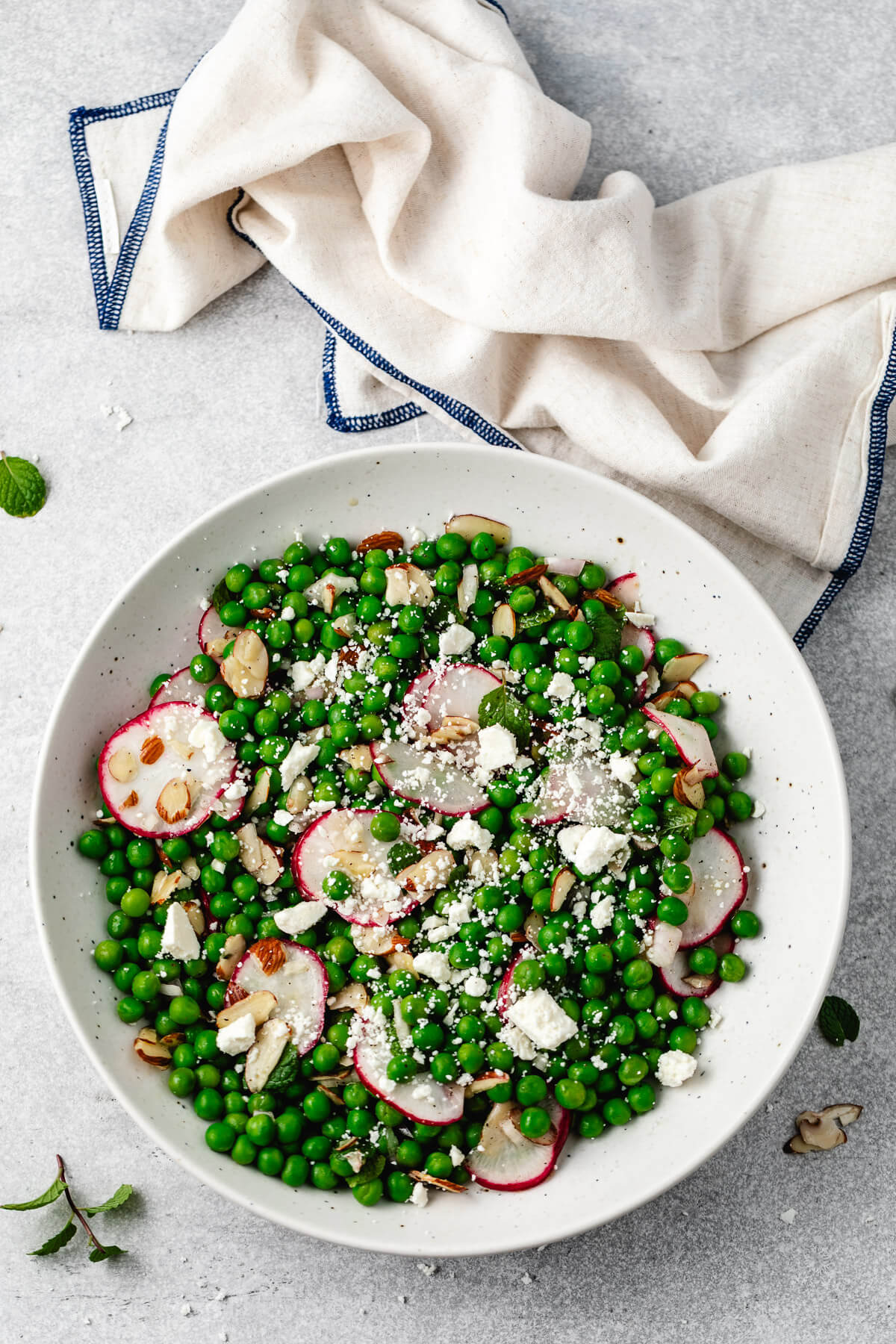 Overhead shot of a bowl of pea salad with feta, radishes, and almonds on a gray background.