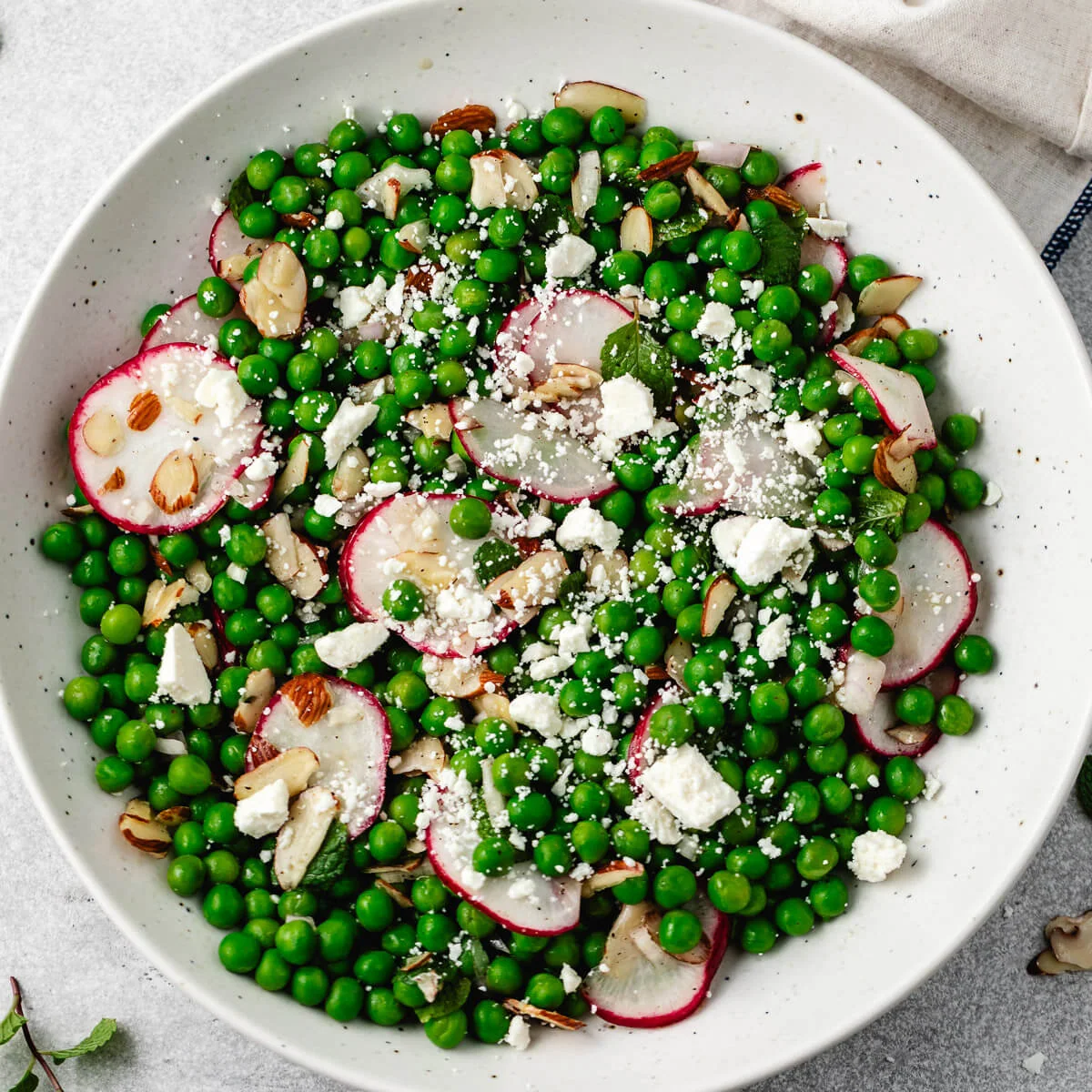 A fresh pea salad with radishes, mint, feta, and toasted almonds in a white bowl.