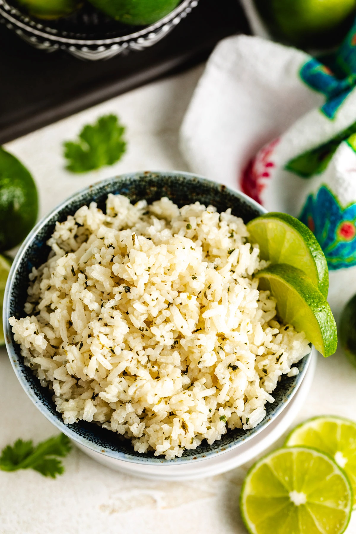 Close-up of rice flecked with herbs in a bowl, surrounded by limes and a cilantro leaf on the counter.