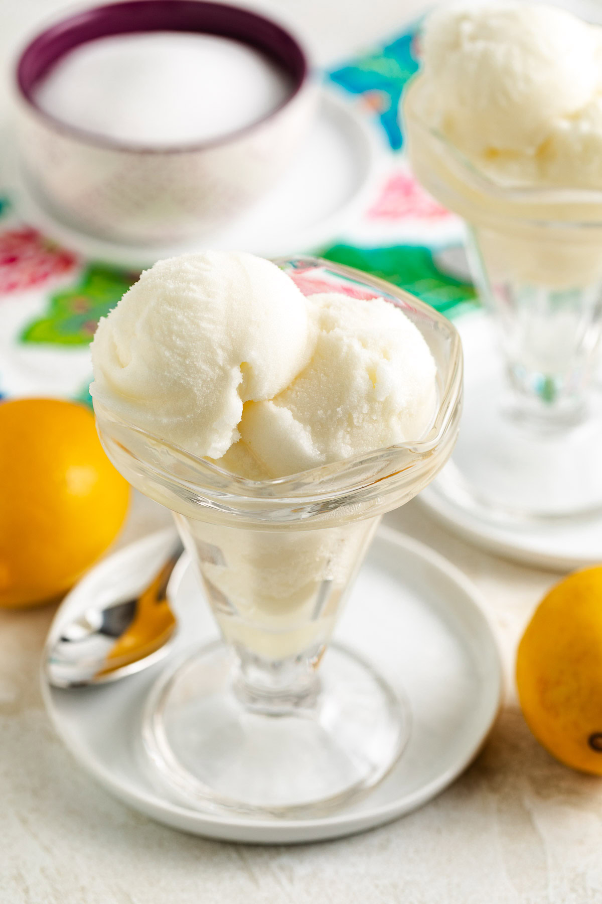 Close-up of a frosty citrus dessert in a glass dish on a plate, with lemons, a spoon, and a patterned cloth.