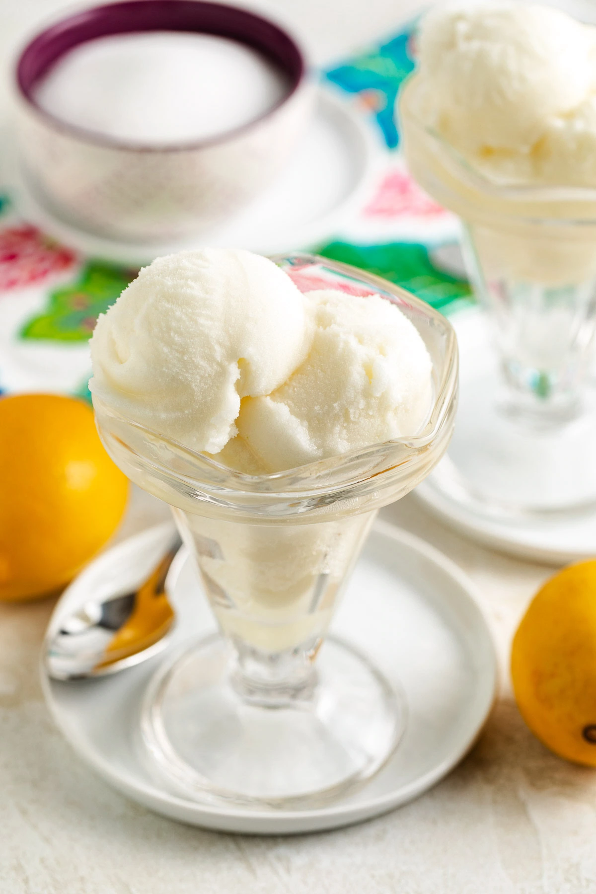 Close-up of a frosty citrus dessert in a glass dish on a plate, with lemons, a spoon, and a patterned cloth.