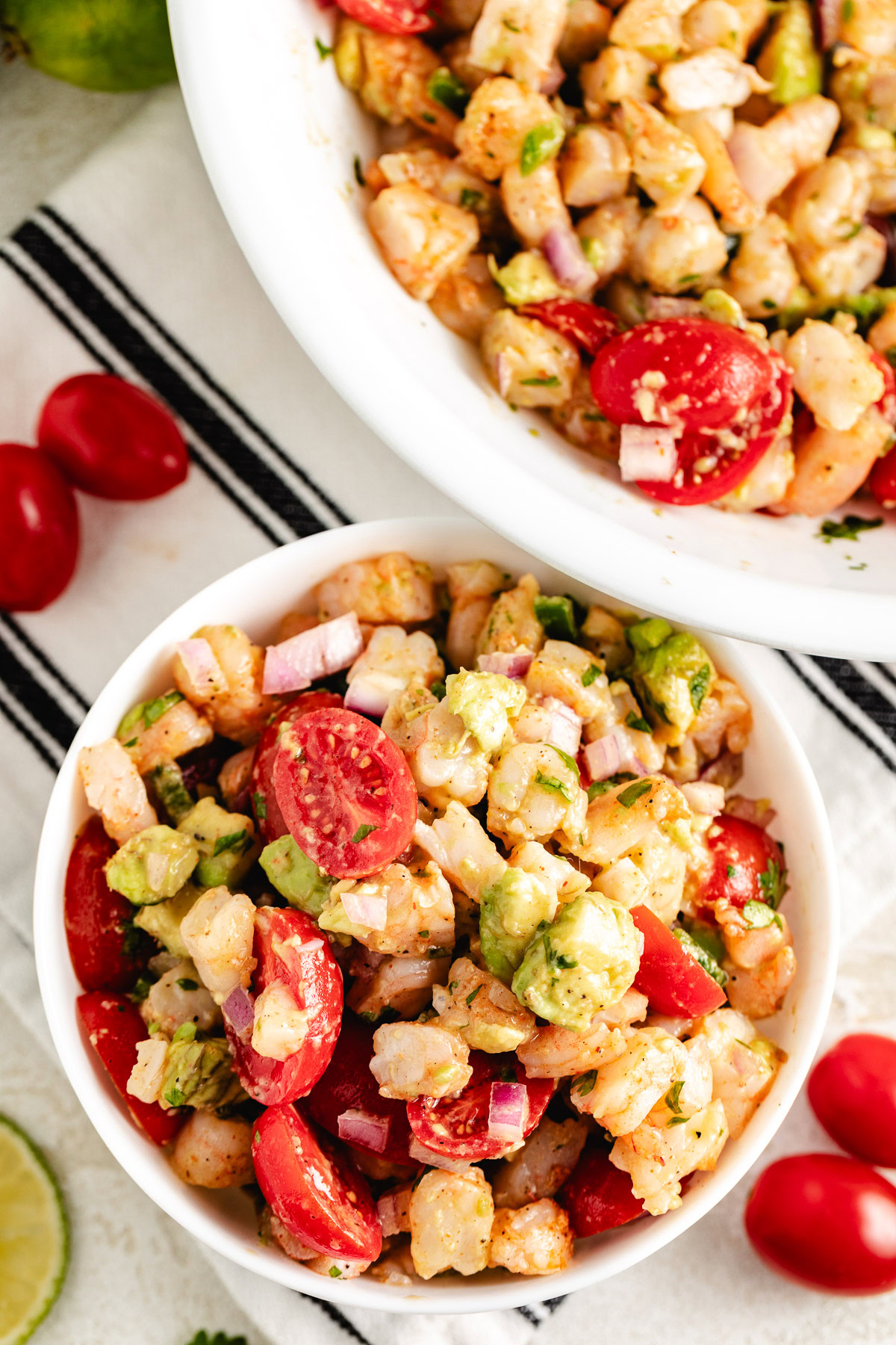 Close-up of lime-dressed shrimp with avocado chunks, sliced cherry tomatoes, and red onion in a small white bowl.
