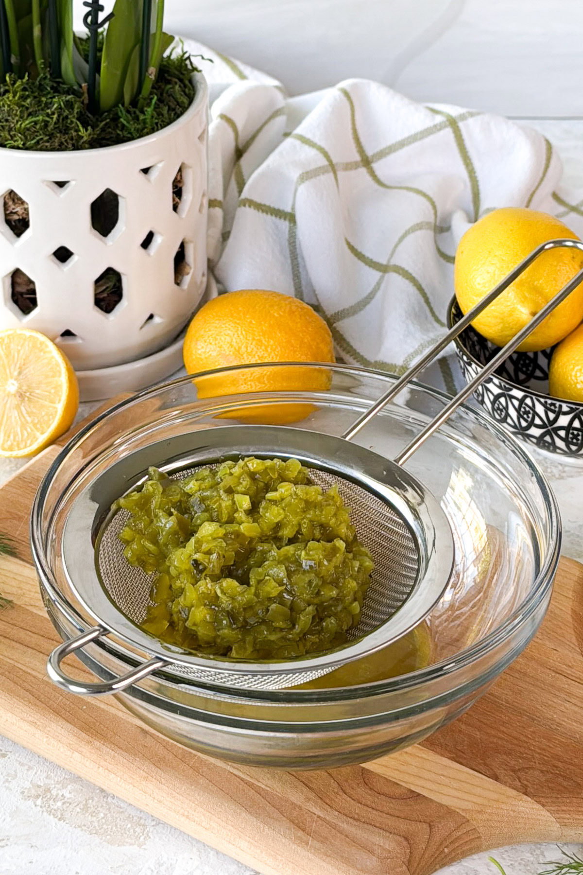 Dill relish draining in a fine-mesh strainer over a glass bowl on a cutting board with lemons nearby.