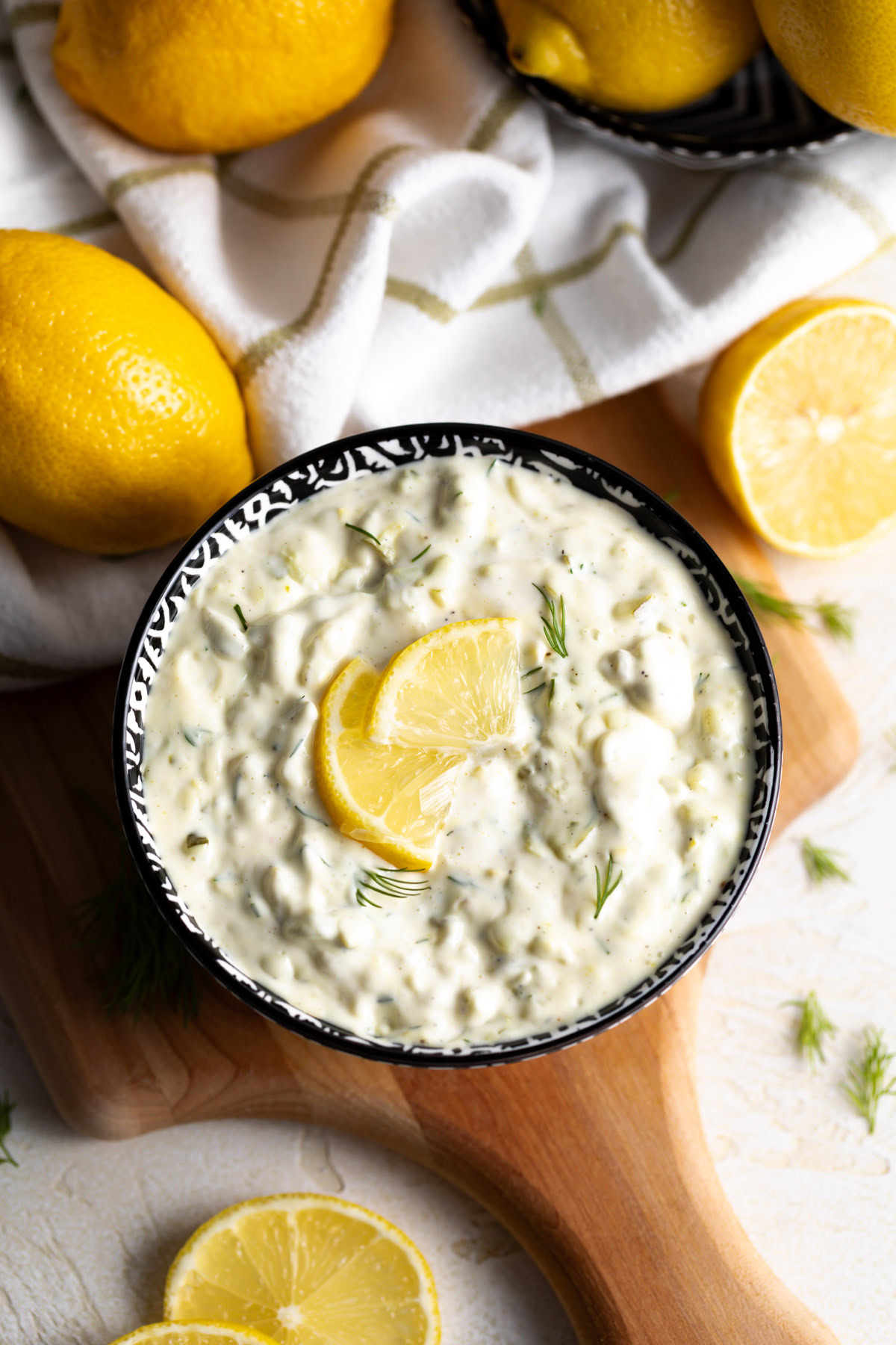Bowl of tartar sauce with a lemon wedge on top, set on a wooden board with whole lemons and a tea towel in the background.
