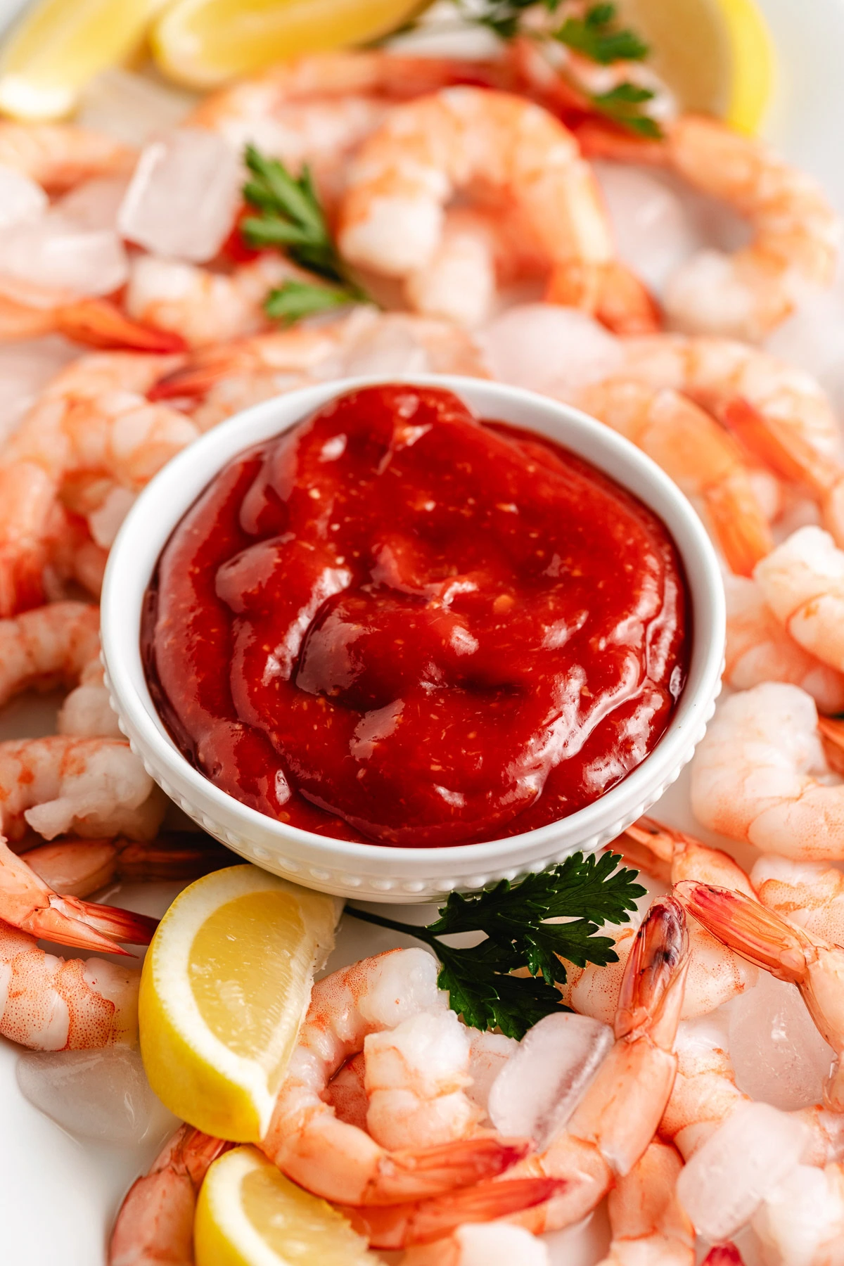 White bowl filled with rich red sauce on a tray of cooked shrimp with lemon slices.