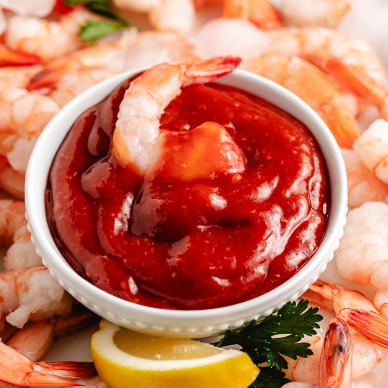 White bowl of shrimp cocktail sauce topped with a shrimp, surrounded by lemon wedges and chilled shrimp.