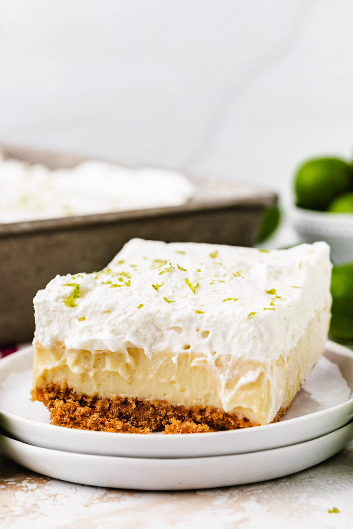 A slice of lime bars on a plate with fresh limes in the background.